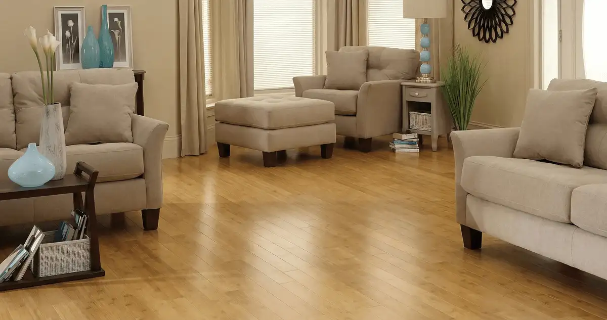 A living room with professionally installed bamboo flooring, beige sofas, a matching ottoman, a side table with a blue lamp, vases, framed pictures, and large windows with blinds and curtains in Syracuse, NY